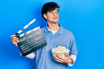 Handsome hipster young man eating popcorn holding cinema clapboard smiling looking to the side and...