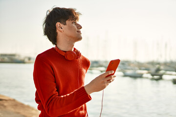 Young hispanic man smiling happy using smartphone and headphones at the beach.
