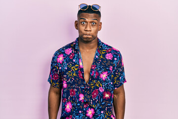 Young black man wearing hawaiian shirt and sunglasses puffing cheeks with funny face. mouth inflated with air, crazy expression.