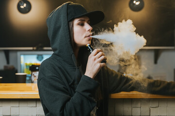 Young pretty woman in cap smoke an electronic cigarette at the vape shop. Hip-hop style. Closeup. - 480805678