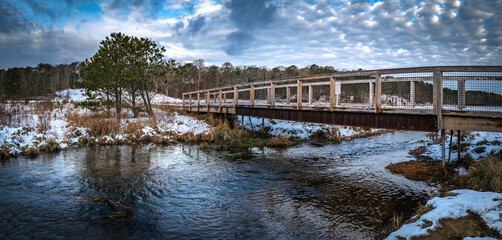 Fototapeta na wymiar Wooden bridge over the river in the snow-covered pine tree forest and grassy wilderness under dramatic cumulous clouds on Cape Cod, Massachusetts.