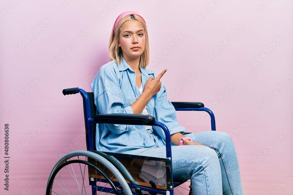 Sticker Beautiful blonde woman sitting on wheelchair pointing with hand finger to the side showing advertisement, serious and calm face