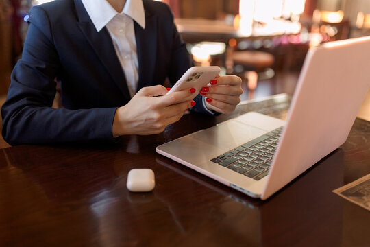 Close-up. Hands Of A Woman With Red Manicure Working With Laptop And Phone. There Are Wireless Headphones On The Table Nearby.