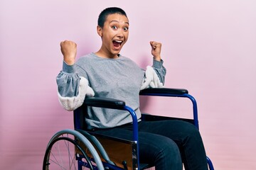 Beautiful hispanic woman with short hair sitting on wheelchair celebrating surprised and amazed for...