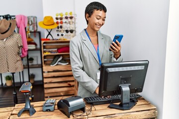 Young hispanic woman shopkeeper smiling confident using smartphone at clothing store