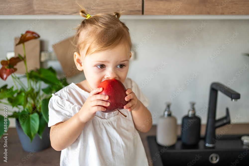 Wall mural Baby girl blonde eating an apple in the kitchen, concept of healthy food for children