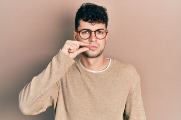 Young hispanic man wearing casual clothes and glasses mouth and lips shut as zip with fingers....
