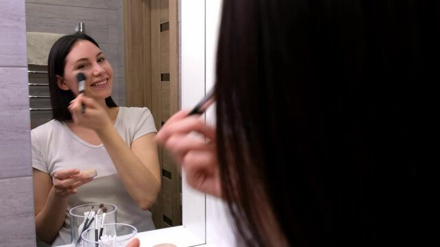 A beautiful young brunette woman looks in the mirror, applies powder with a brush on her face in the bathroom and smiles. Cosmetics and self-care