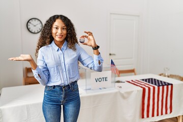 Beautiful hispanic woman standing by at political campaign by voting ballot smiling cheerful presenting and pointing with palm of hand looking at the camera.