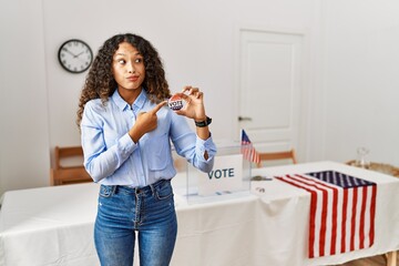 Beautiful hispanic woman standing by at political campaign by voting ballot pointing aside worried...