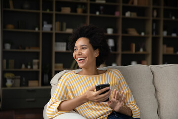Happy candid young attractive African American woman using cellphone, looking in distance laughing at funny message, watching entertaining photo video content online, relaxing on comfortable sofa.