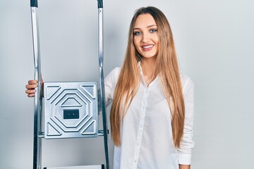 Young blonde girl holding ladder looking positive and happy standing and smiling with a confident smile showing teeth