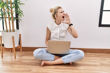 Young blonde woman using computer laptop sitting on the floor at the living room shouting and screaming loud to side with hand on mouth. communication concept.