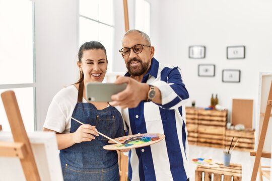 Young artist woman and senior painter man at art studio classroom painting on canvas with brush and oil painting taking a selfie picture with smartphone
