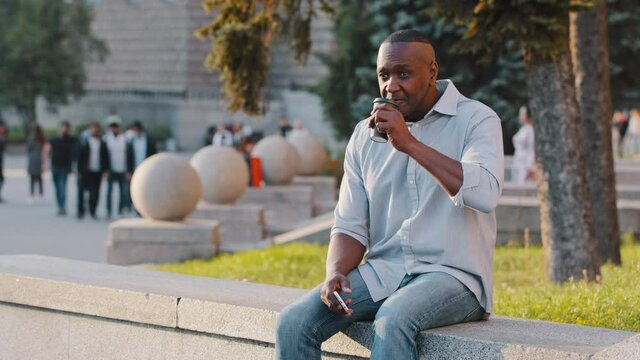 Adult mature american businessman holding smartphone in hand talking partner on speakerphone outdoors. African man chatting during break enjoys aromatic hot drink of coffee or tea in disposable cup
