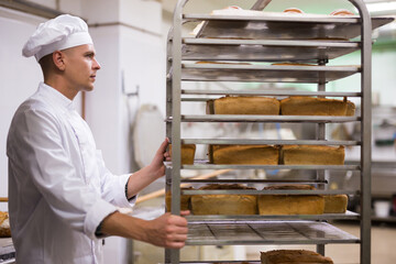 Portrait of confident male chef pushing rack trolley with fresh bread in bakery kitchen