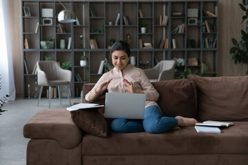 Happy young beautiful Indian woman in wired earphones holding video call in computer, communicating with teacher distantly learning foreign language, sitting on cozy sofa at home.