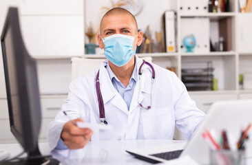 Doctor in protective medical mask sitting at workplace with computer in her office