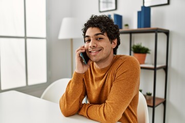 Young hispanic man talking on the smartphone sitting on the table at home.
