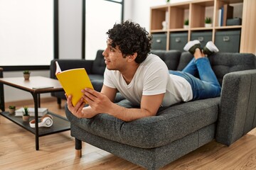 Young hispanic man reading book lying on the sofa at home.