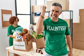 Middle age man wearing volunteer t shirt at donations stand angry and mad raising fist frustrated and furious while shouting with anger. rage and aggressive concept.