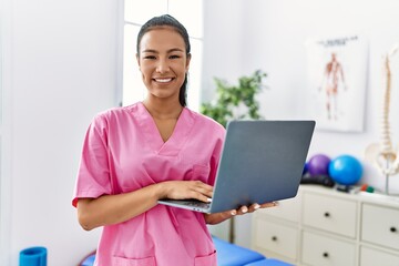 Young hispanic physiotherapist woman using computer laptop at medical clinic looking positive and happy standing and smiling with a confident smile showing teeth