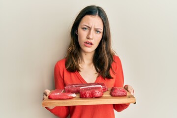 Young brunette woman holding board with raw meat in shock face, looking skeptical and sarcastic,...