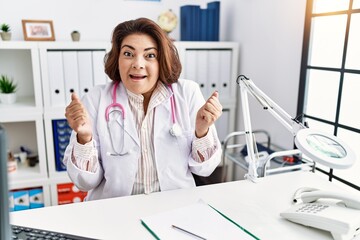Obraz na płótnie Canvas Middle age hispanic woman wearing doctor uniform and stethoscope at the clinic celebrating surprised and amazed for success with arms raised and open eyes. winner concept.