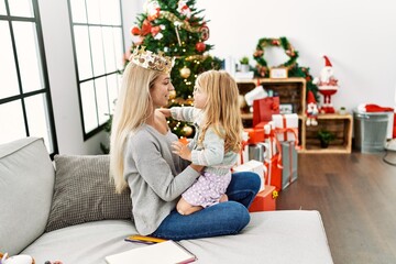 Mother and daughter wearing queen cronw hugging each other sitting by christmas tree at home