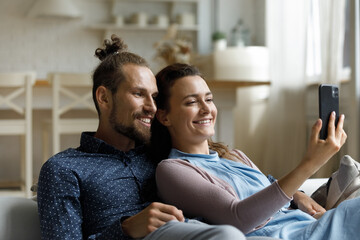 Fototapeta premium Joyful affectionate young family couple looking at smartphone screen, holding pleasant web camera call conversation or watching funny video online in social network, sitting on cozy couch at home.