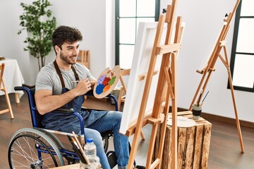 Young hispanic artist disabled man smiling happy painting at art studio.