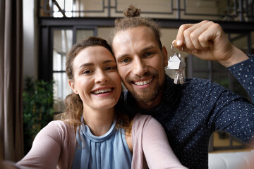 Happy loving bonding young married family couple looking at camera showing keys from own apartment, streaming stories in social network or posing for selfie photo, celebrating moving into new dwelling