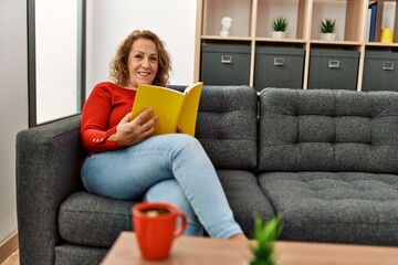 Middle age caucasian woman reading book sitting on the sofa at home.