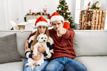Young couple of wife and husband sitting on the sofa wearing christmas hat at home pointing with finger to the camera and to you, confident gesture looking serious