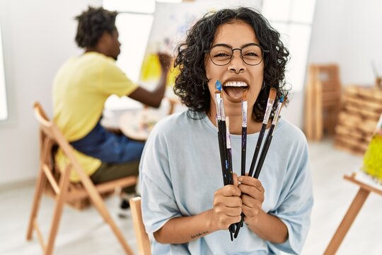 African american artist couple smiling happy painting at art studio. Woman holding paintbrushes sitting on chair.