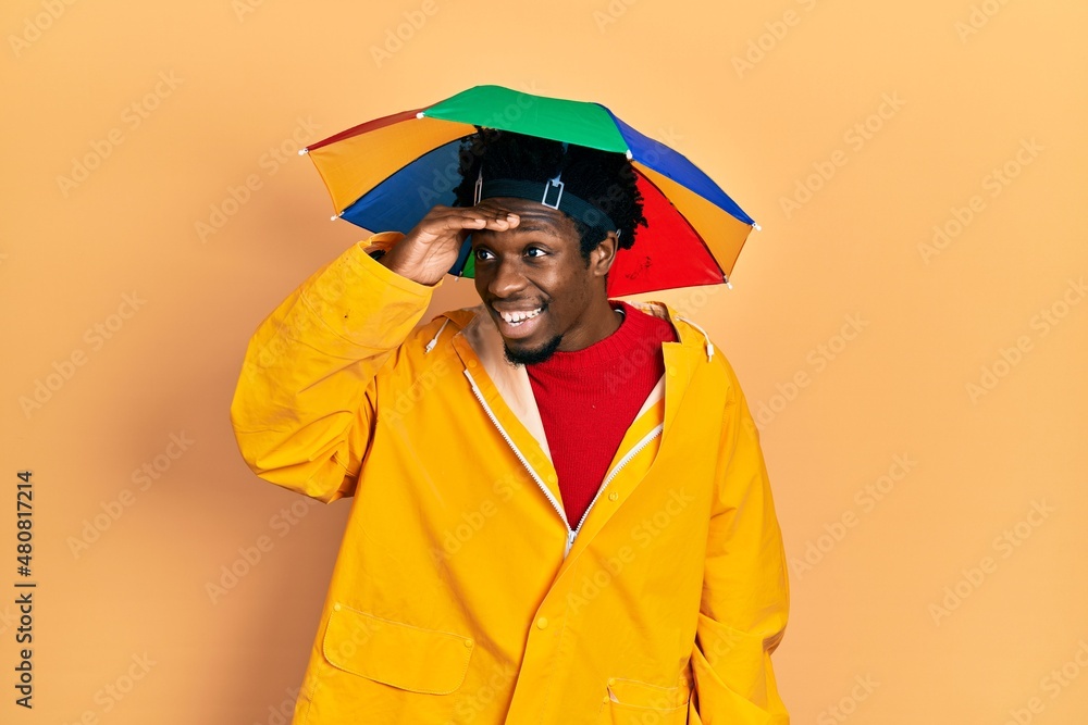 Wall mural Young african american man wearing yellow raincoat very happy and smiling looking far away with hand over head. searching concept.