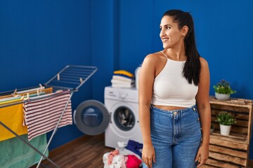 Young hispanic woman at laundry room looking away to side with smile on face, natural expression. laughing confident.