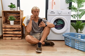 Young blond man doing laundry using smartphone mouth and lips shut as zip with fingers. secret and...