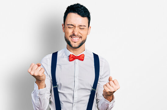 Hispanic man with beard wearing hipster look with bow tie and suspenders very happy and excited doing winner gesture with arms raised, smiling and screaming for success. celebration concept.
