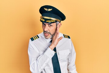 Handsome middle age man with grey hair wearing airplane pilot uniform hand on mouth telling secret rumor, whispering malicious talk conversation