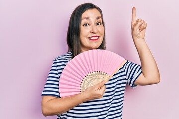 Middle age hispanic woman waving hand fan cooling air in summer smiling with an idea or question pointing finger with happy face, number one