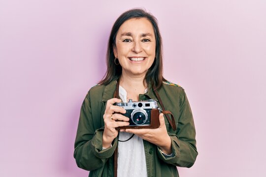 Middle age hispanic woman holding vintage camera looking positive and happy standing and smiling with a confident smile showing teeth