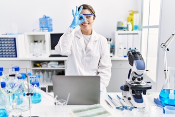 Young hispanic woman wearing scientist uniform working at laboratory smiling happy doing ok sign with hand on eye looking through fingers