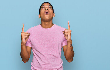 Young handsome hispanic man wearing casual pink t shirt amazed and surprised looking up and...