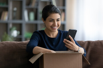 Happy beautiful young Indian woman unboxing carton parcel at home, checking order in cellphone...