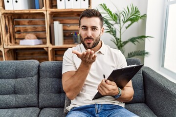 Handsome hispanic man holding clipboard working at psychology clinic looking at the camera blowing a kiss with hand on air being lovely and sexy. love expression.