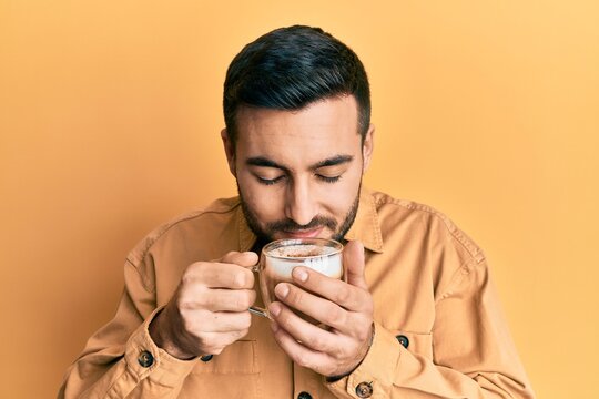 Handsome hispanic man enjoying a cup of coffee over yellow background
