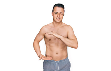 Handsome young man wearing swimwear shirtless gesturing with hands showing big and large size sign, measure symbol. smiling looking at the camera. measuring concept.