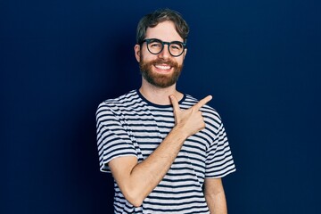 Caucasian man with beard wearing striped t shirt and glasses cheerful with a smile of face pointing with hand and finger up to the side with happy and natural expression on face