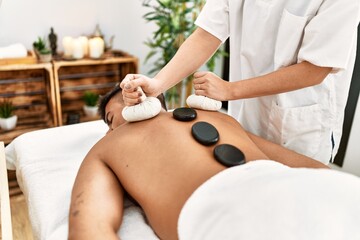 Obraz premium Young hispanic man relaxed having back massage using black stones and thai bags at beauty center